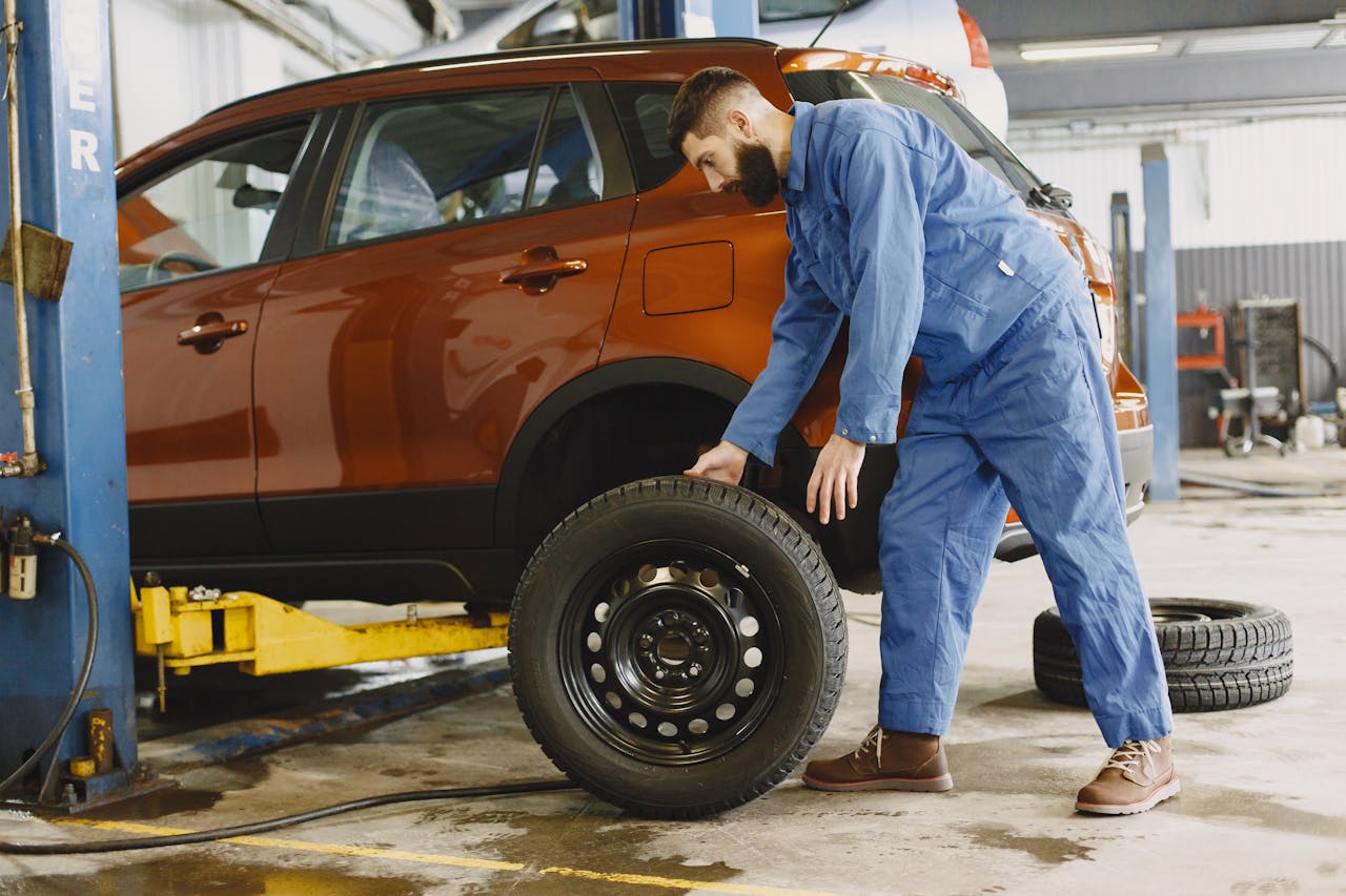 Home Mechanic in blue coveralls replacing tire on red SUV in repair shop garage.